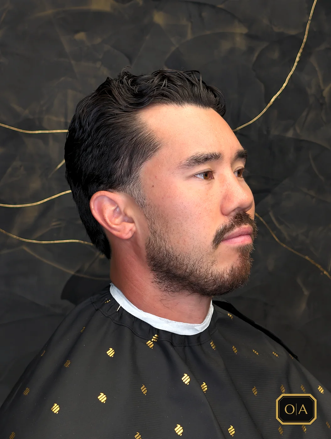 A finished full taper with longer hair on top slicked back loosely, seen from the side against the shop's black wall