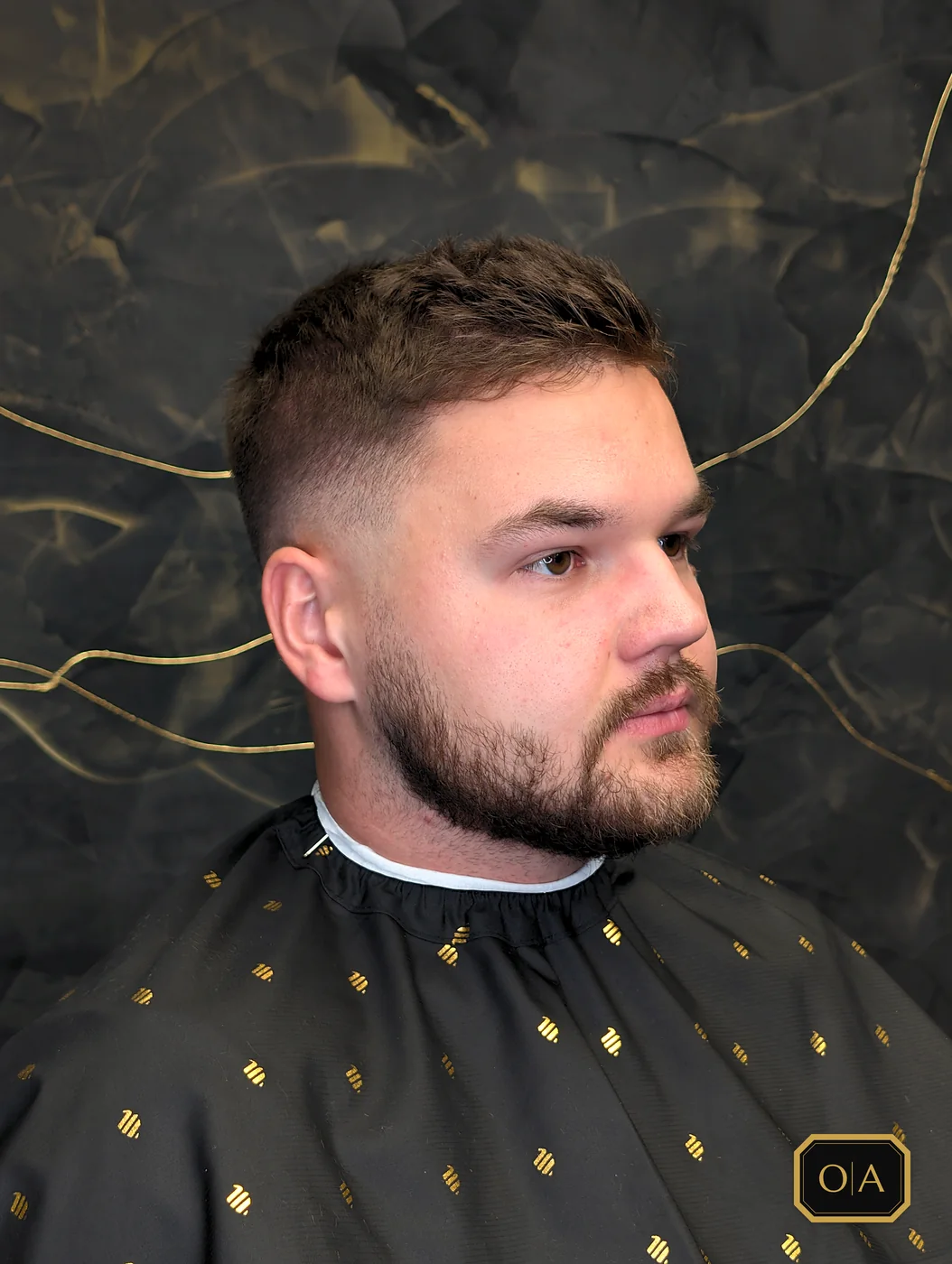 A finished mid bald fade with short textured hair on top swept up and over at the front, seen from the side against the shop's black wall