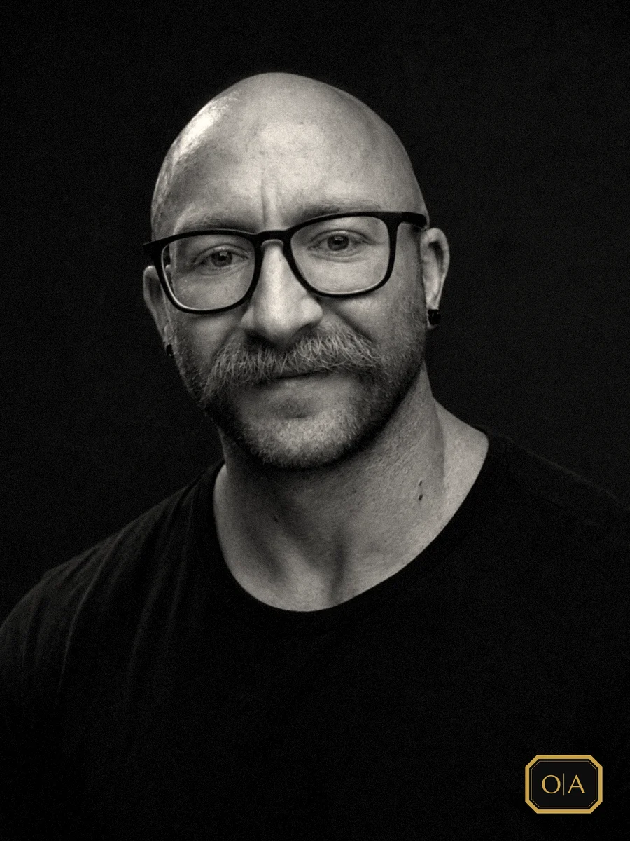 Gus, the barber behind Onyx & Aurum, in glasses and a black T-shirt, in black and white