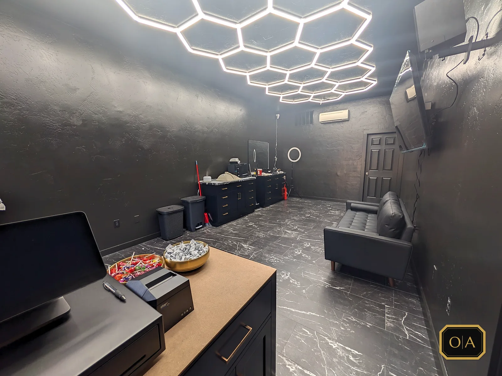 The finished front room from behind the checkout counter: the hexagon light grid overhead, black marble-look floor, two black vanities with brass pulls along the far wall and a black leather couch by the wall-mounted TV.