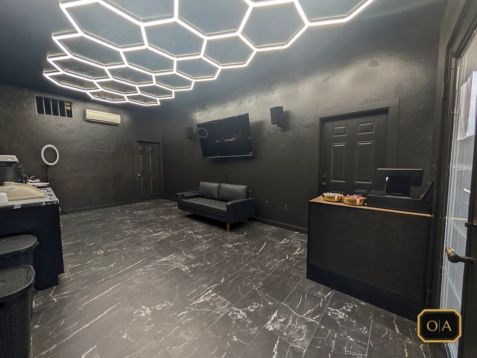 The front room from the entrance: the lit hexagon ceiling, a black leather couch under the TV and speakers, and the checkout counter by the front door with bowls of candy on top.
