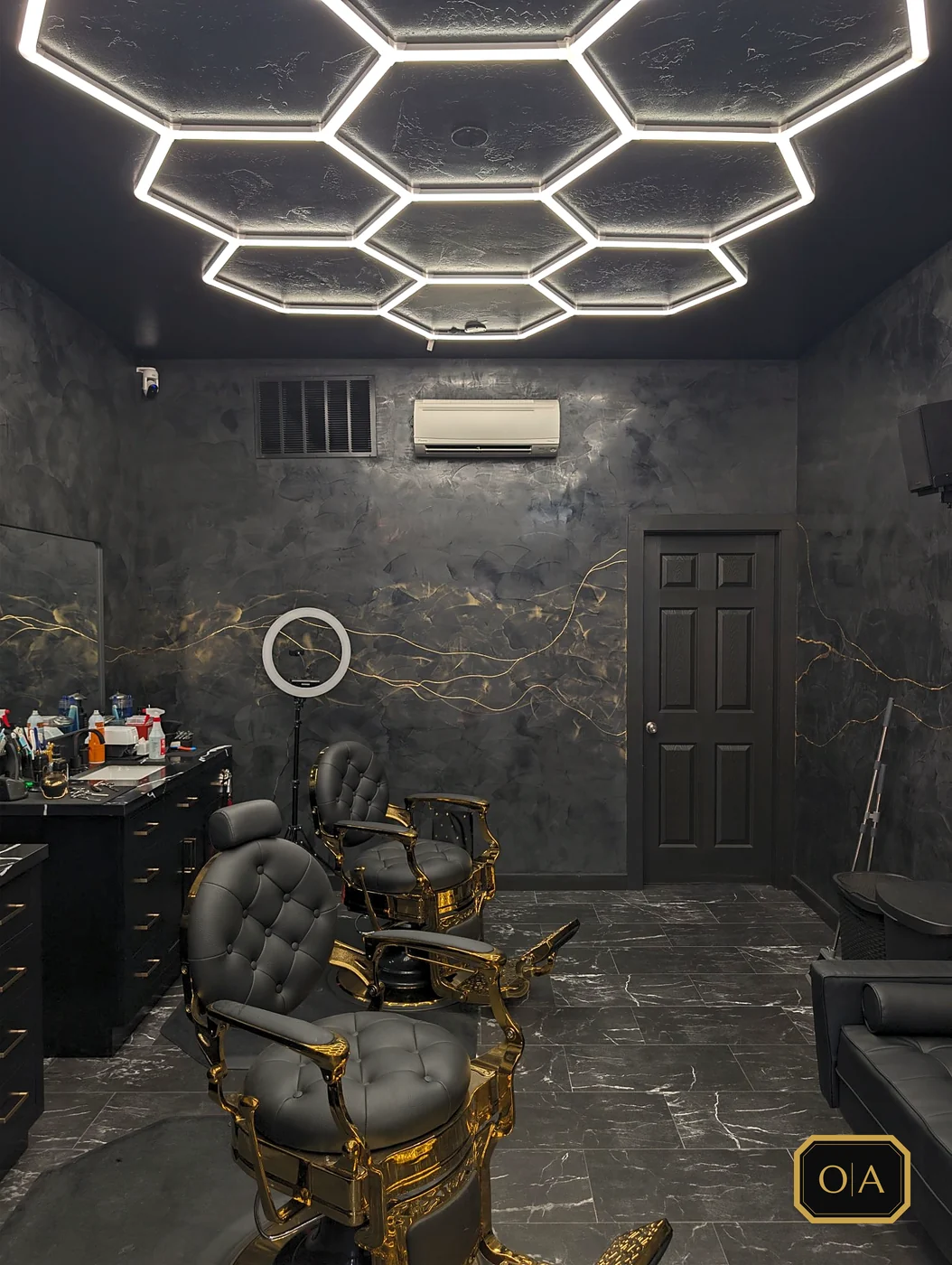 The finished chair room: two gold-and-black barber chairs under the lit hexagon ceiling, black walls with fine gold veins running across them, a black marble-look floor and a black vanity along the left wall.